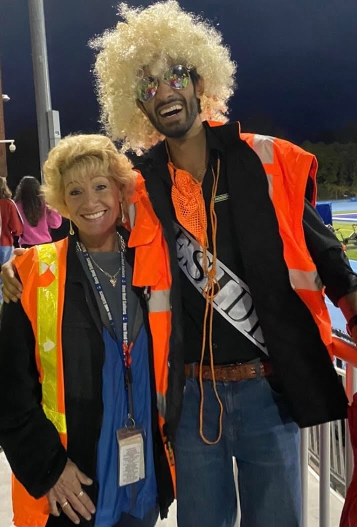 Two people in colorful outfits and wigs pose together at a nighttime event. Both wear bright orange safety vests over their costumes.