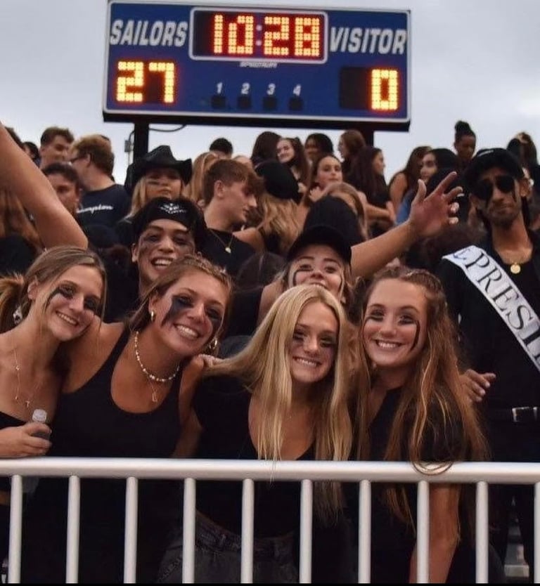 Group of smiling fans watching a high school football game from bleachers with scoreboard showing Sailors leading 27-0 in background