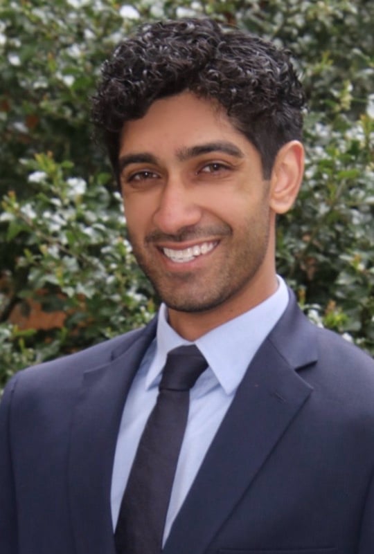 Professional headshot of a smiling man wearing a dark blazer and dark tie against a leafy green background
